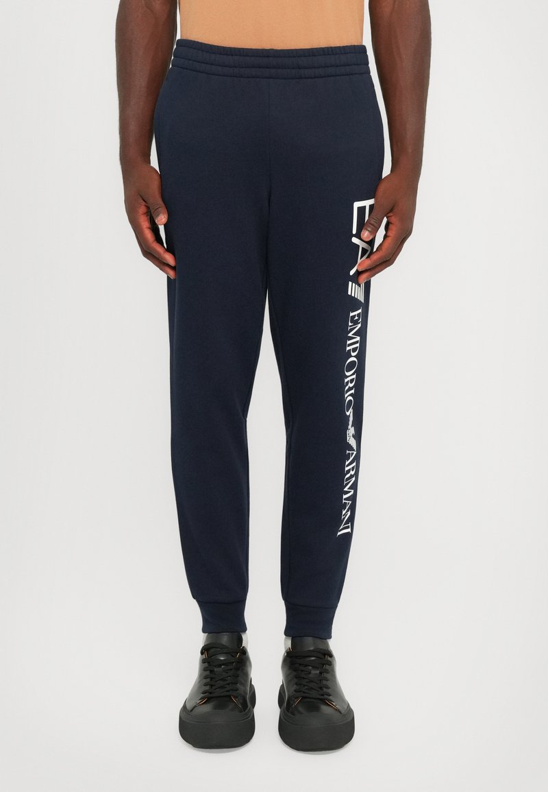 Navy sweatpants with elastic waistband, tapered legs, and white branding along the side. Paired with black sneakers featuring thick soles.