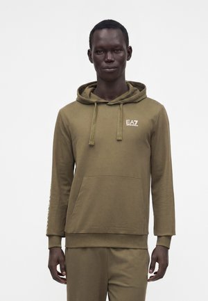 Άνδρας που φοράει ελιά EA7 Emporio Armani φούτερ με κουκούλα και ταιριαστό παντελόνι, στέκεται μπροστά σε ένα απλό ανοιχτό φόντο.