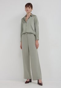 Blouse en satin vert clair avec un col, boutonnière à l'avant et manches longues, associée à un pantalon large assorti. Des chaussures à talons sombres complètent le look.