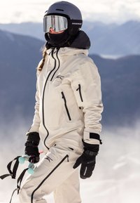 White waterproof ski suit with black accents, zippers, and gloves. Large black helmet and goggles. Snowy mountain background.