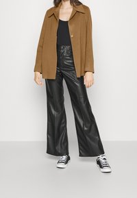 Hnědá oversized bunda z vlněné směsi, černý top, kalhoty z umělé kůže s vysokým pasem a černobílé tenisky se šněrováním.
