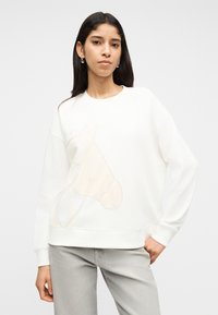 Γυναίκα με μακριά σκούρα μαλλιά που φοράει ασημένια σκουλαρίκια κρίκους, άσπρο φούτερ με μπεζ σχέδιο αλόγου και ανοιχτόχρωμα γκρι τζιν, ποζάροντας με το χέρι στην τσέπη.