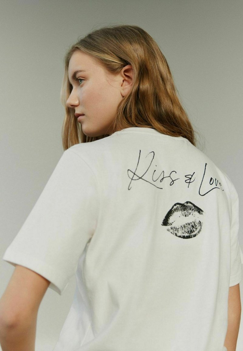 Valkoinen puuvillapaita, jossa on musta suudelma painatus ja "Kiss & Love" -teksti takana; lyhyet hihat, rento malli, sileä rakenne.