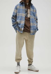 Blauw en beige geruite flanellen shirt over een bruine hoodie, beige joggingbroek en witte sneakers met grijze sokken.