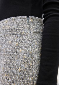 Κλείσιμο ενός ατόμου που φοράει υφασμάτινη γκρι και μπεζ καρό φούστα με κρυφό φερμουάρ, σε συνδυασμό με μαύρο μακρυμάνικο τοπ.