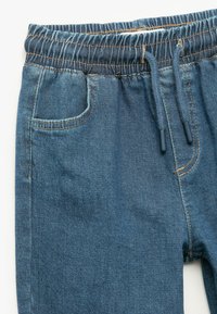 Pants de denim azul con cintura elástica y cordón de ajuste, con bolsillos frontales y detalles de costura en naranja.