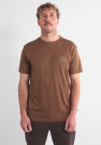 T-shirt en coton marron avec un col rond, des manches courtes, et un petit motif graphique avant comprenant du texte et un design circulaire.