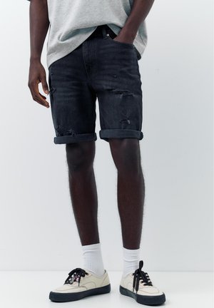 Homme portant un short en jean noir déchiré retroussé, des chaussettes blanches et des baskets écru, debout avec une main dans la poche.