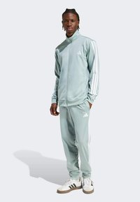 Svijetlozeleni trenirka s bijelim Adidas prugama na rukavima i hlačama. Glatka, atletska tkanina; jakna s patentnim zatvaračem; logo na prsima i bedru.