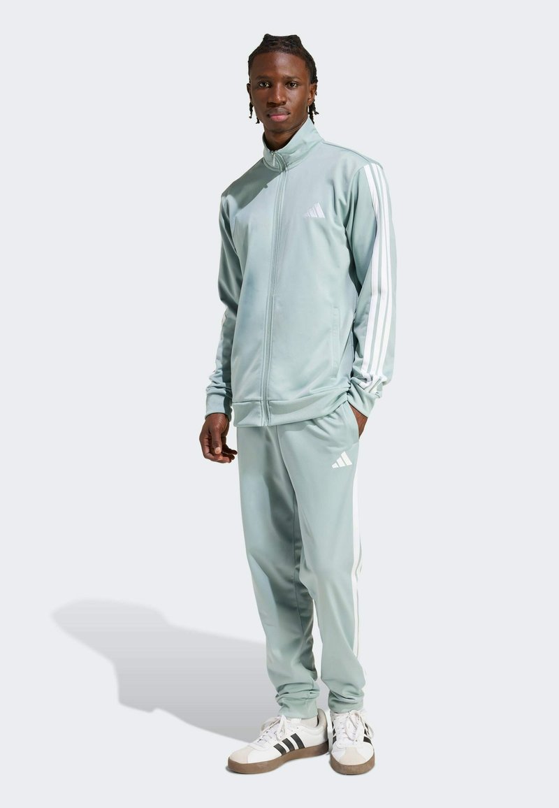 Svijetlozeleni trenirka s bijelim Adidas prugama na rukavima i hlačama. Glatka, atletska tkanina; jakna s patentnim zatvaračem; logo na prsima i bedru.