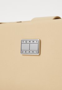 Bolso sintético beige con una textura suave que presenta una placa rectangular de logo en plata grabada con "TOMMY JEANS."