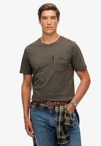 Oliwkowa koszulka z krótkim rękawem z kieszonką na piersi. Nosić ją z niebieskimi dżinsami i koszulą w kratę zawiązaną w talii. Brązowy skórzany pasek i zegarek.