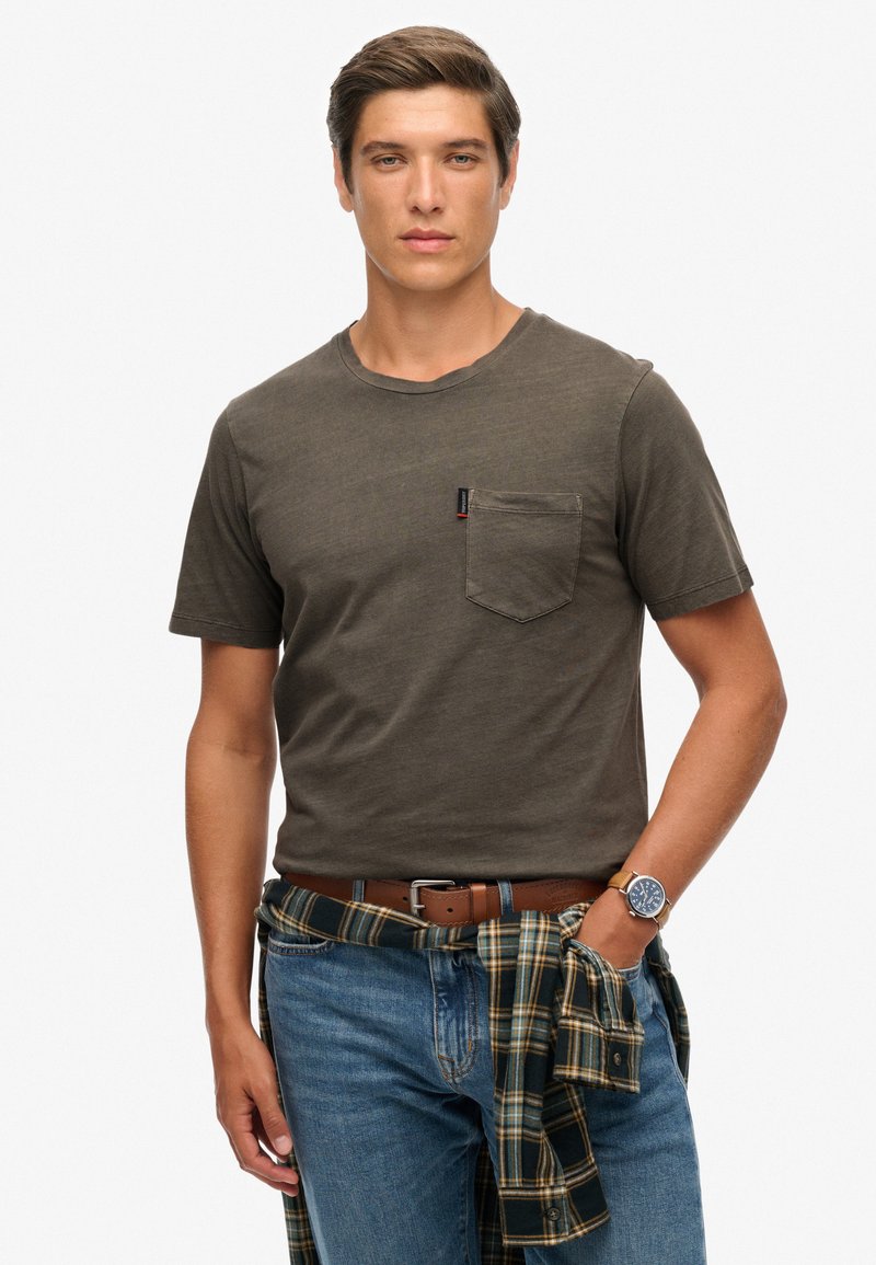 Oliwkowa koszulka z krótkim rękawem z kieszonką na piersi. Nosić ją z niebieskimi dżinsami i koszulą w kratę zawiązaną w talii. Brązowy skórzany pasek i zegarek.