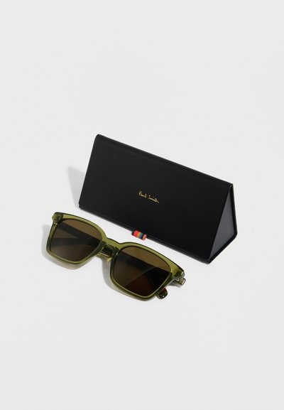 Gafas de sol verdes con lentes marrones y montura gruesa. Acompañadas de un estuche triangular negro con logotipo dorado y un detalle a rayas.