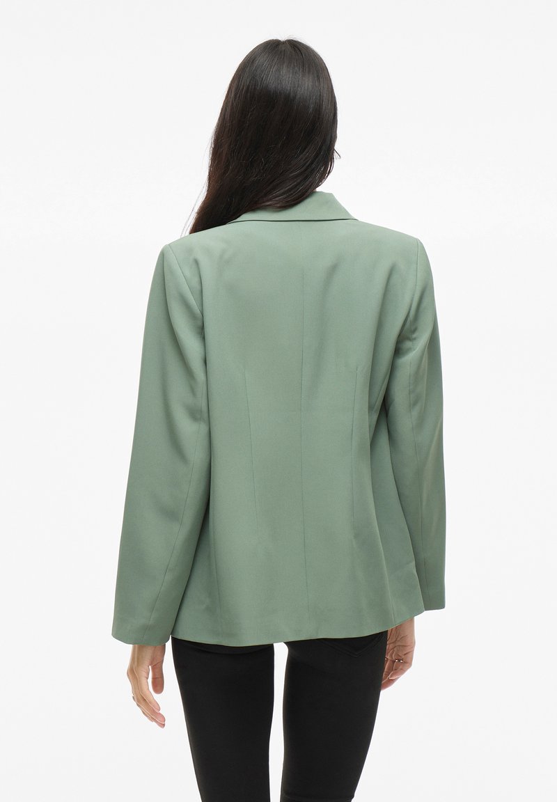 Blazer vert à manches longues, coupe ajustée, revers crantés et texture lisse. Vue de l'arrière, montrant des lignes épurées et un minimum de détails.