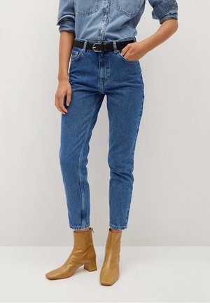 Woman wearing high-waist blue jeans with a black belt, a rolled-up denim shirt, and tan ankle boots with block heels.