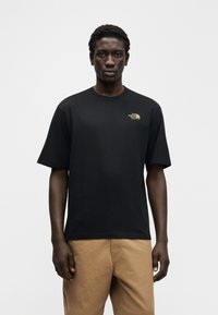 Μαύρο βαμβακερό μπλουζάκι με κοντό μανίκι και στρογγυλή λαιμόκοψη, με ένα μικρό χρυσό λογότυπο στο επάνω αριστερό μέρος του στήθους.