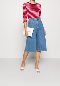Rot-weißer gestreifter Langarmshirt, kombiniert mit hochgeschnittenen blauen Jeans-Culottes und einer weißen Clutch. Weiße Sandalen mit offenem Zehen und Absatz.
