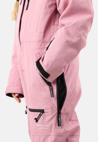 Tuta da sci rosa con vestibilità ampia, dotata di una ventilazione con zip, molteplici tasche, polsini regolabili e un logo sul davanti. Texture liscia.