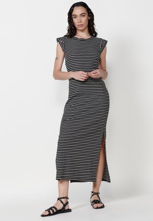 Robe maxi rayée noire et blanche avec des manches courtes, fente sur le côté, design ajusté, accompagnée de sandales noires à lanières.