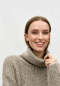 Hnědo-krémový pruhovaný rolák s měkkou texturou, oversized střihem a žebrovanými detaily. Model se usmívá a drží látku blízko krku.