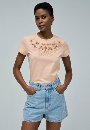 Camiseta de melocotón claro con diseño de cut-out floral en el pecho, mangas cortas, combinada con pantalones cortos de mezclilla de cintura alta en azul claro.