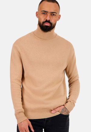 Pull à col roulé beige en tricot côtelé avec une silhouette ajustée. Manches longues et col haut. Associé à un pantalon noir.