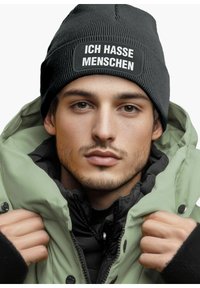 Black knitted beanie with white text patch "ICH HASSE MENSCHEN," worn with a zippered green puffer jacket over a black layer.