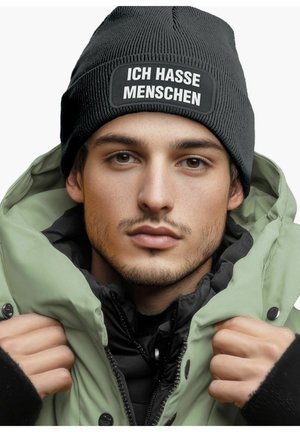 Black knitted beanie with white text patch "ICH HASSE MENSCHEN," worn with a zippered green puffer jacket over a black layer.