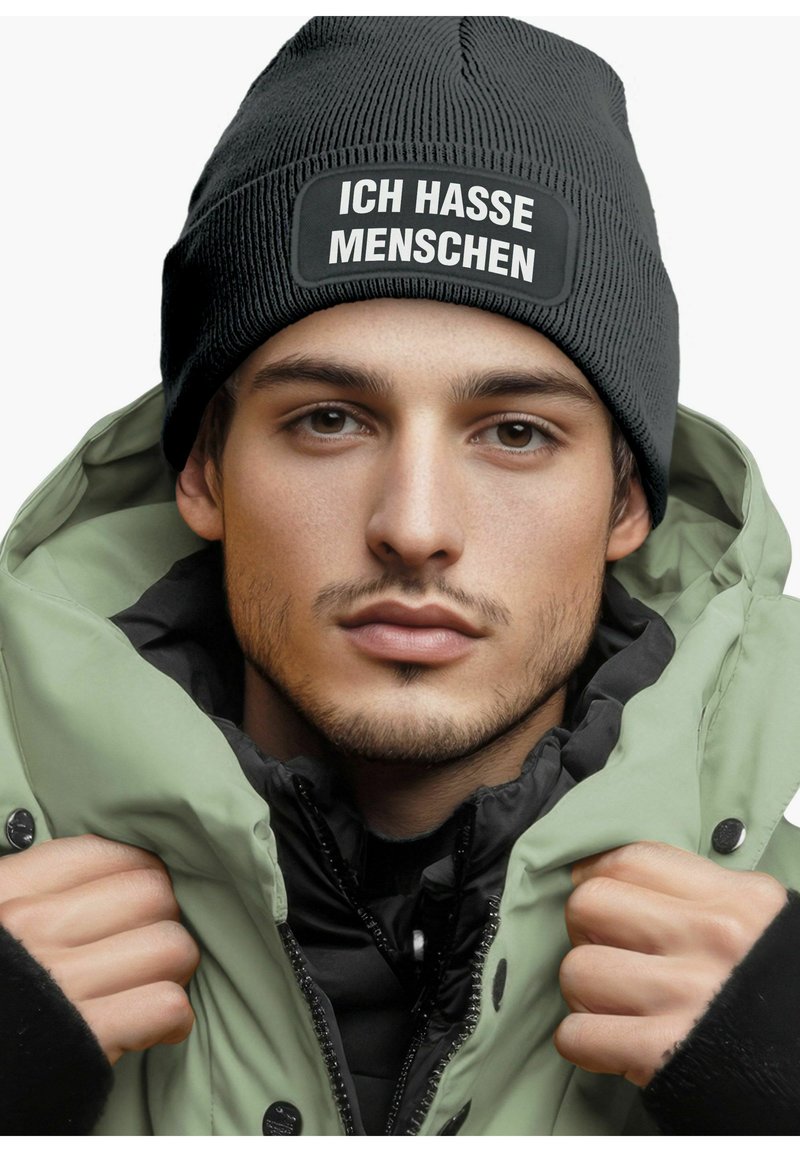 Black knitted beanie with white text patch "ICH HASSE MENSCHEN," worn with a zippered green puffer jacket over a black layer.