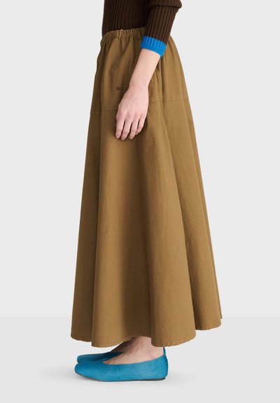 Personne debout portant une jupe longue marron avec une taille élastique, des ballerines bleues et un pull marron avec des poignets bleus, sur un fond uni.