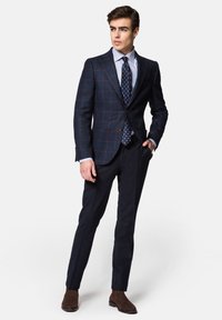 Granatowa marynarka w krate z brązowymi akcentami, zestawiona z jasną koszulą w paski i krawatem w kropki. Spodnie slim-fit oraz brązowe botki za kostkę.