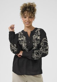 Sort bluse med V-hals, der har detaljeret hvid blomsterbroderi på ærmerne og kroppen. Langærmede med knapmanchetter.