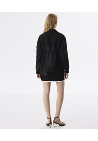 Chaqueta negra acolchada con textura en canalé, que presenta una cintura elástica y un corte holgado. Combinada con una falda corta blanca y sandalias negras.