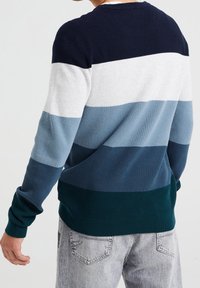 Hombre vistiendo un suéter de punto waffle de manga larga con bloques de color horizontales en navy, blanco, azul claro, azul oscuro y verde azulado, combinado con jeans de lavado claro.