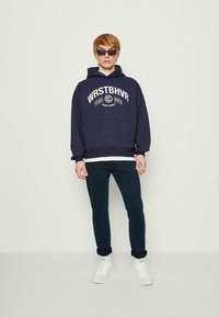 Giovane che indossa occhiali da sole neri, felpa blu navy con stampa bianca, pantaloni scuri e sneakers bianche, in piedi davanti a uno sfondo chiaro e uniforme.