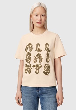 Jeune femme aux cheveux blonds platine lisses porte un t-shirt beige avec des lettres brunes ornées formant le mot "ALANIS" et un jean bleu.