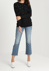 Svart tröja med texturerad yta och pärlliknande detaljer, kombinerad med ljusblå cropped jeans och vita klackar.