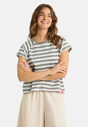 Junge Frau mit welligem Haar, die lächelt, trägt ein grün-weiß gestreiftes Kurzarmshirt und helle, beige, lockere Hosen, Arme verschränkt.