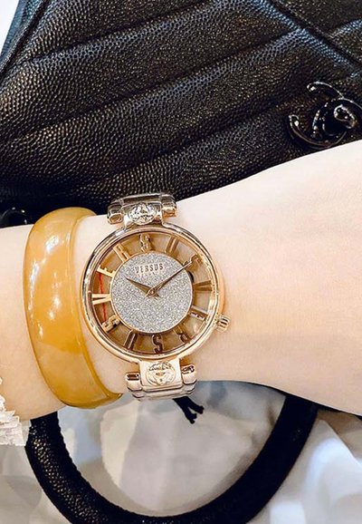 Gold-tone watch with a round case, silver glitter dial, and clear numeral accents. Accompanied by a yellow bangle and textured black bag.