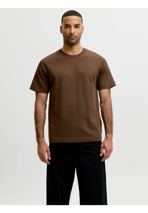 Mand stående med front mod, iført en ensfarvet brun kortærmet t-shirt og sorte bukser mod en ensfarvet lys baggrund.