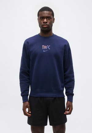 Mann trägt dunkelblauen Sweatshirt mit „THFC“ und Nike-Logo, kombiniert mit schwarzen Shorts, steht vor schlicht hellem Hintergrund.