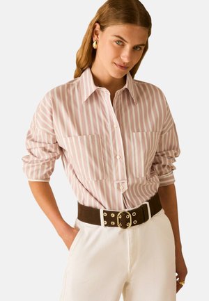 Vrouw draagt een roze-wit gestreept overhemd met knopen en opgerolde mouwen, witte broek, zwarte riem met gouden gesp en gouden oorbellen in hoepelvorm.