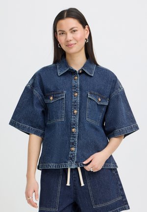 Ung kvinde med langt mørkt hår iført mørk denim skjorte med korte ærmer og knapper samt matchende shorts med hvidt snørrebånd.