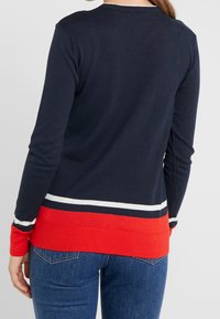Kobieta w granatowym swetrze z białymi i czerwonymi paskami przy mankietach i u dołu, w połączeniu z niebieskimi dżinsami, pokazana od tyłu.