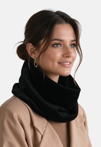 Mujer con el cabello recogido lleva un abrigo beige y una bufanda negra de lazo, sonriendo ligeramente, con pendientes dorados colgantes sobre un fondo blanco.