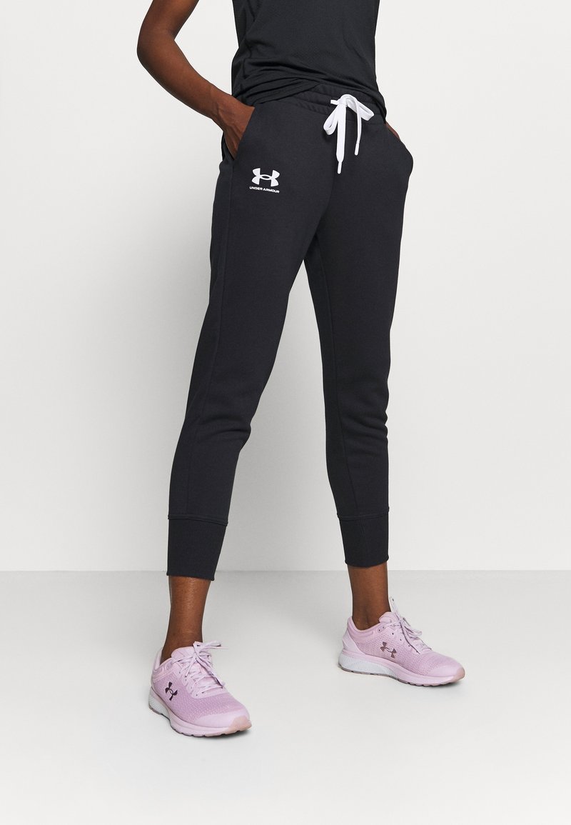 Persoon in zwarte Under Armour joggers met een witte trekkoord en roze Under Armour hardloopschoenen, staand met de handen in de zakken.