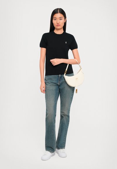 Woman in black short-sleeve top, blue jeans, and white sneakers holding a white crescent-shaped handbag against a plain background.