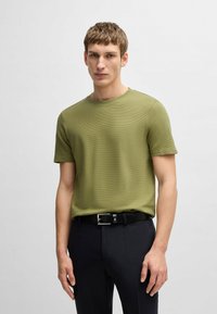 T-shirt verde oliva con texture, colletto rotondo e maniche corte, abbinata a pantaloni neri e una cintura nera con fibbia in argento.