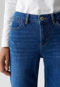Gros plan sur une personne portant un jean en denim bleu et une chemise à manches longues blanche, montrant la taille à l'avant et la main gauche détendue sur le côté.
